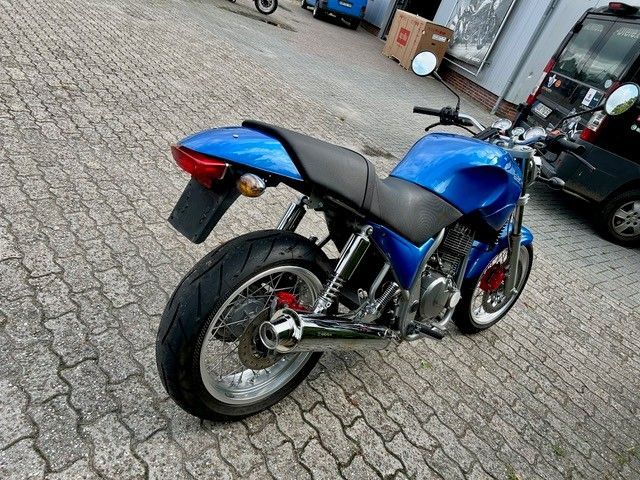 Auf Kopfsteinpflaster geparktes blaues Motorrad mit individuellem Heck und Speichenrädern.