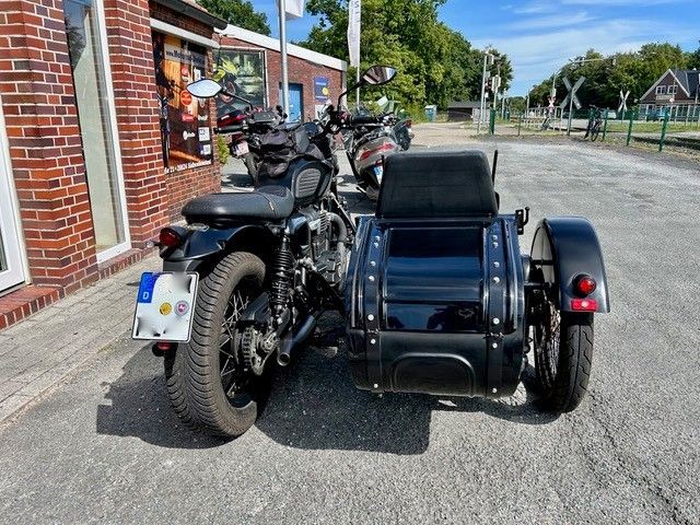 Schwarzes Motorrad mit Beiwagen auf einem gepflasterten Platz geparkt. Im Hintergrund ist ein rotes Backsteingebäude zu sehen.