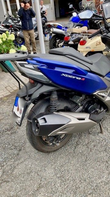 Ein blauer Roller parkt auf Schotter, im Hintergrund sind Menschen und andere Roller zu sehen.
