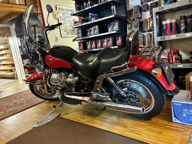 Ein rotes Motorrad steht drinnen auf einem Teppich neben Regalen und Werkzeugen in einer Garage oder Werkstatt.