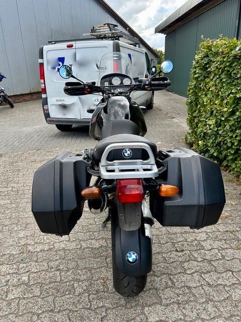 Rückansicht eines schwarzen BMW-Motorrads mit Satteltaschen, das auf Kopfsteinpflaster neben einem Lieferwagen geparkt ist.