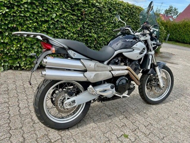 Ein silbernes Tourenmotorrad parkt auf einer gepflasterten Auffahrt neben einer grünen Hecke.