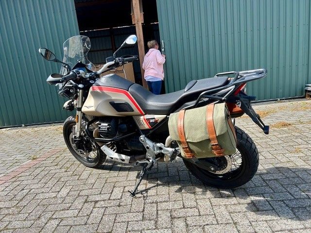 Ein Motorrad mit Seitentasche parkt auf einem gepflasterten Weg vor einer grünen Mauer; eine Person steht in der Nähe.