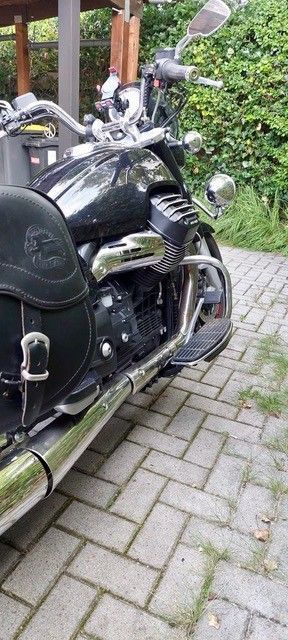 Schwarzes Motorrad auf Ziegelpflaster geparkt. Chromdetails, schwarze Satteltasche und grünes Laub im Hintergrund.