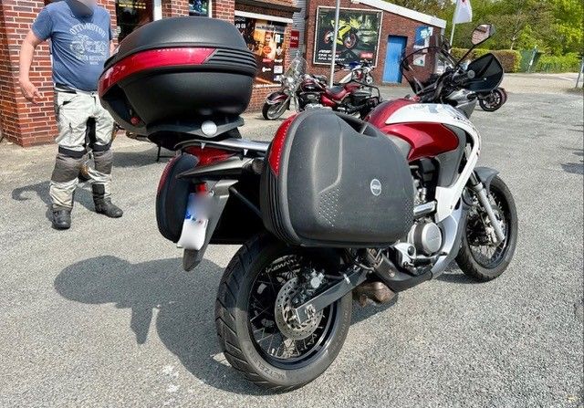 Draußen geparktes Motorrad mit roten und schwarzen Packtaschen und Topcase. Fahrer in blauem Hemd und grauer Hose.