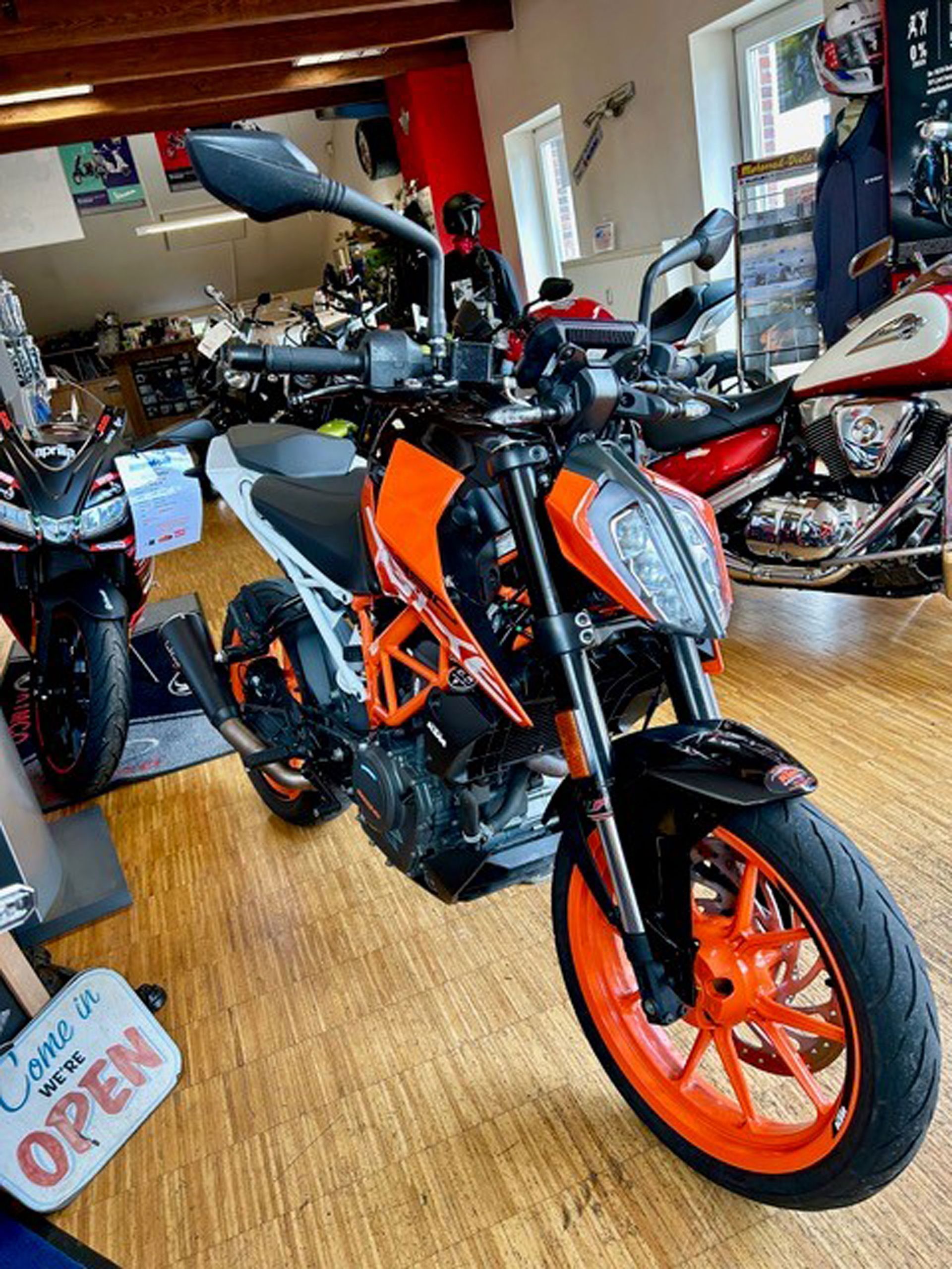 Orange-schwarzes KTM Duke-Motorrad in einem Ausstellungsraum, mit orangefarbenen Rädern und Rahmenakzenten. Das Schild „Kommen Sie herein, wir haben GEÖFFNET“ ist sichtbar.