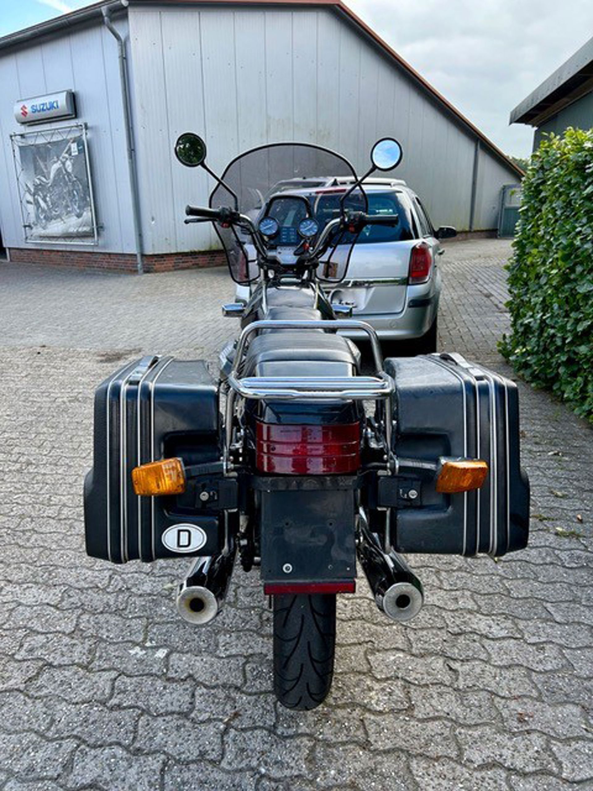 Ein schwarzes Motorrad mit Packtaschen, geparkt auf einer gemauerten Fläche vor einem Gebäude und einem silbernen Auto.