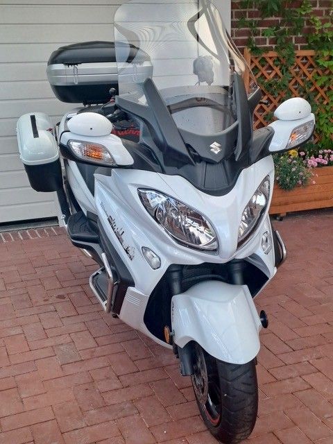 Weißer Suzuki Burgman-Roller, geparkt auf einer gepflasterten Auffahrt. Zur Ausstattung gehören eine Windschutzscheibe, Seitenspiegel und Ablagefächer.