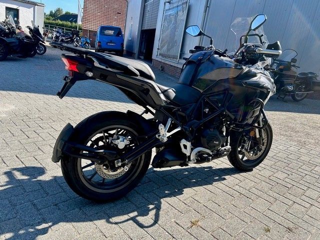 Ein schwarzes Sportmotorrad steht auf einem gepflasterten Parkplatz vor einem Gebäude; im Hintergrund sind weitere Motorräder zu sehen.