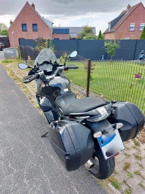 Schwarzes Motorrad mit Satteltaschen, geparkt auf einem Bürgersteig in der Nähe eines Rasenhofs und Backsteinhäusern.
