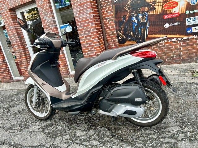 Ein silber-schwarzer Roller parkt auf einem gepflasterten Gehweg vor einem Ladenlokal.