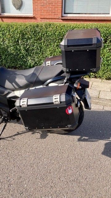 Schwarzes Motorrad mit seitlichen und oberen Aufbewahrungsboxen, auf dem Bürgersteig in der Nähe einer grünen Hecke geparkt.