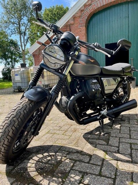 Schwarzes Motorrad Moto Guzzi V7 III Stone, geparkt vor einer grünen Tür und einem grünen Gebäude.