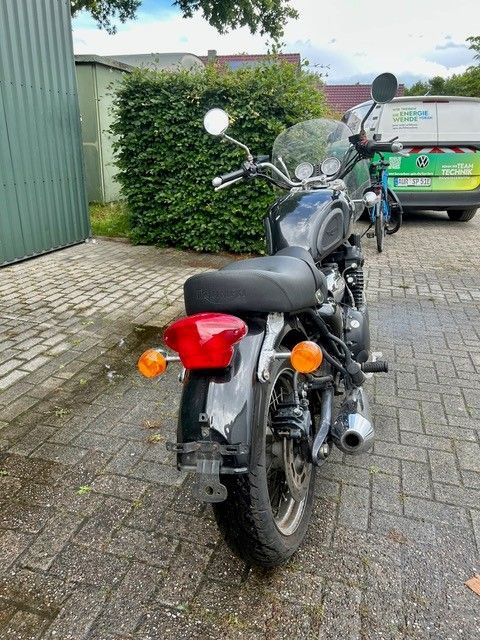 Ein schwarzes Motorrad parkte auf nassem Ziegelpflaster neben einer Hecke und einem grünen Lieferwagen.