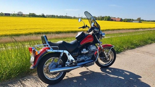 Ein rotes Motorrad parkt auf einer Straße, im Hintergrund ein gelbes Feld unter blauem Himmel.