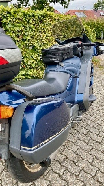 Blaues Motorrad mit Satteltaschen und Topcase, geparkt auf einer Ziegelstraße in der Nähe einer Hecke.
