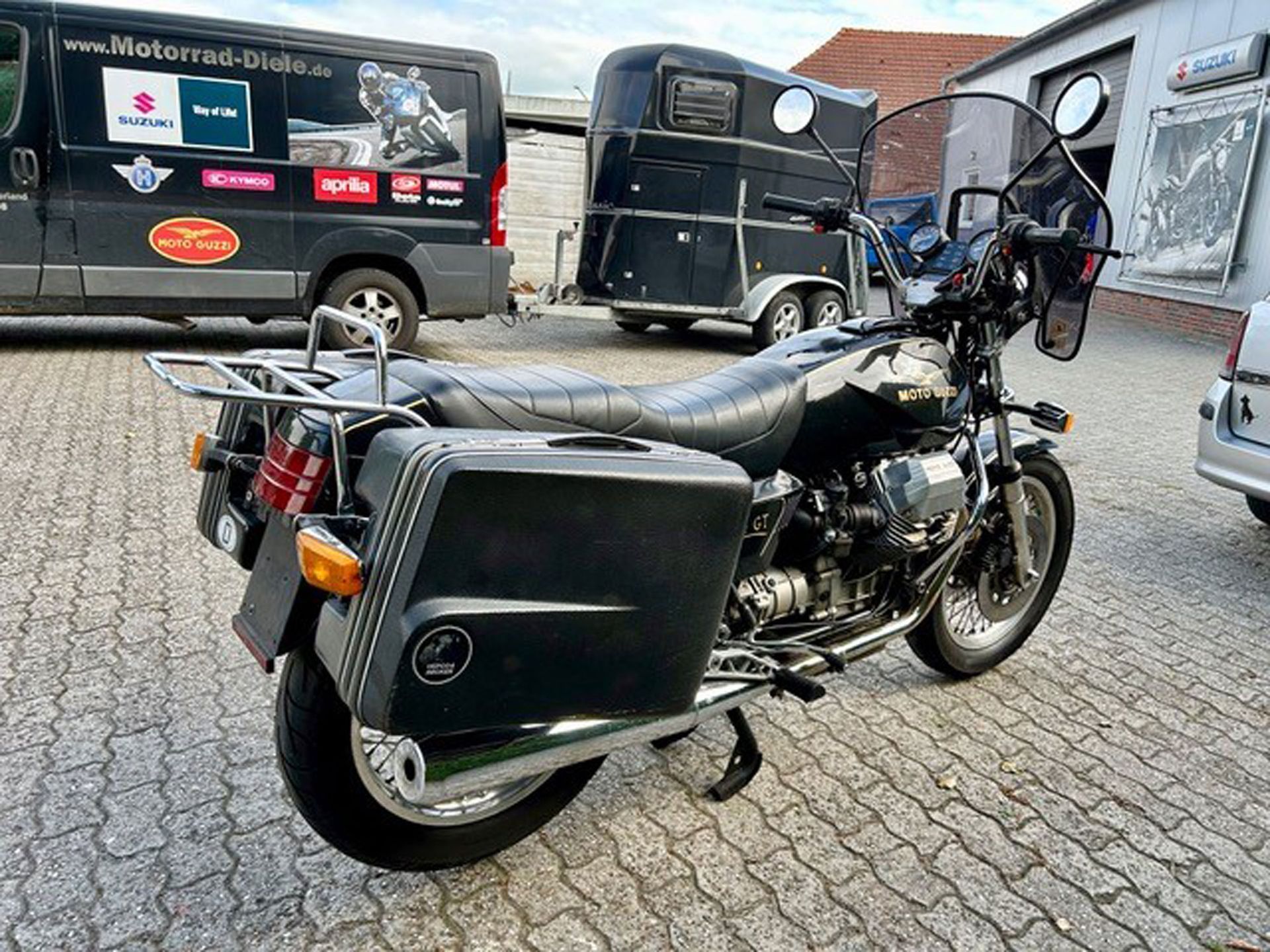 Schwarzes Motorrad mit Satteltaschen auf dem Bürgersteig geparkt, im Hintergrund ein schwarzer Lieferwagen und ein Pferdeanhänger.