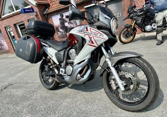 Ein weiß-rotes Honda Transalp-Motorrad mit Seitengepäck ist auf einer befestigten Fläche geparkt; im Hintergrund ist ein weiteres Motorrad zu sehen.