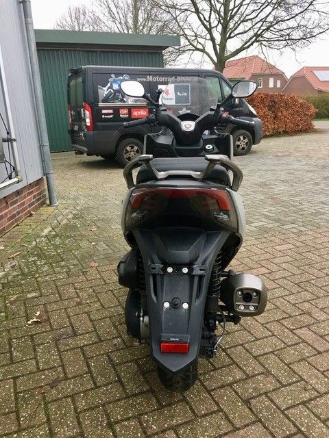 Auf einer gepflasterten Auffahrt ist neben einem Lieferwagen ein Motorroller geparkt.