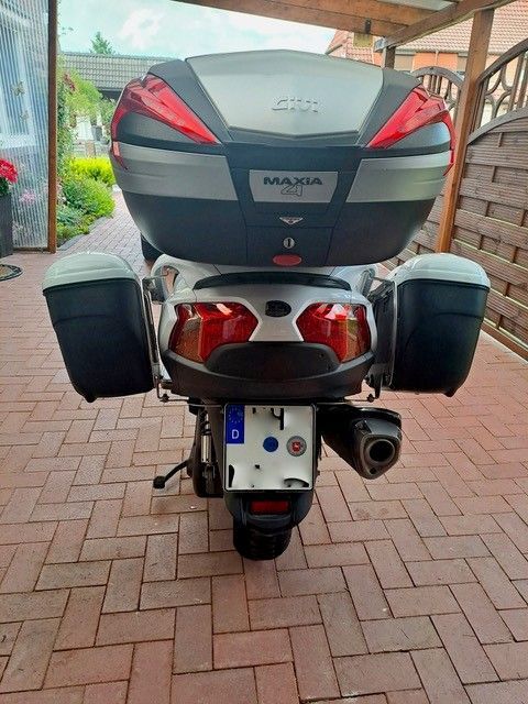 Rückansicht eines weißen Motorrollers mit schwarzen Seitenkoffern und Topcase, der auf einer gemauerten Terrasse geparkt ist.