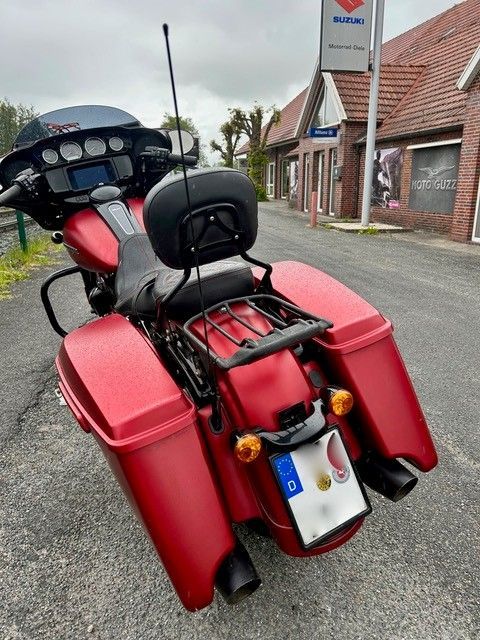 Ein rotes Tourenmotorrad mit Soziussitz und Gepäckträger parkt in einer ruhigen Straße neben Gebäuden.