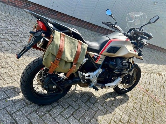 Motorrad mit beiger und schwarzer Lackierung, geparkt auf einer gepflasterten Straße, mit einer grün-braunen Tasche am Heck.