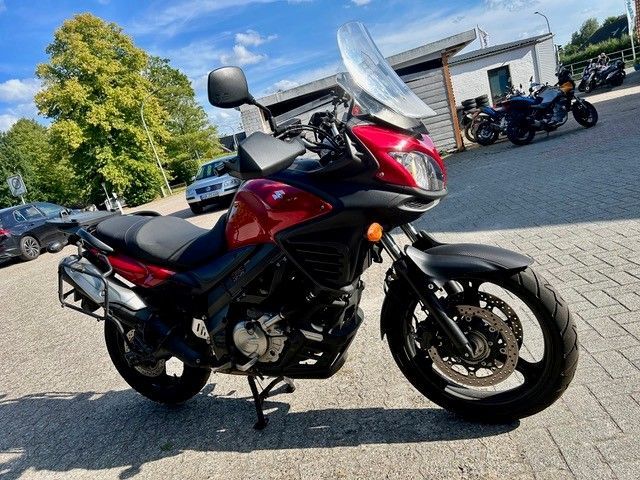 Ein rot-schwarzes Motorrad steht auf einem gepflasterten Parkplatz in der Nähe eines Gebäudes; im Hintergrund sind weitere Motorräder zu sehen.