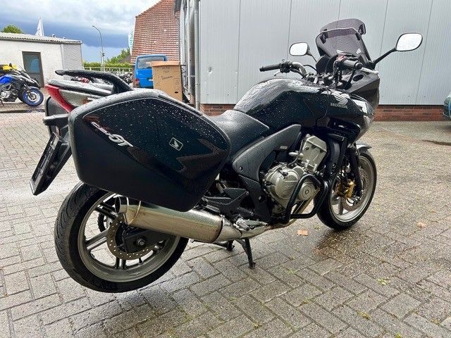 Schwarzes Tourenmotorrad, geparkt auf einem gepflasterten Parkplatz neben einem Gebäude, Ansicht von hinten links