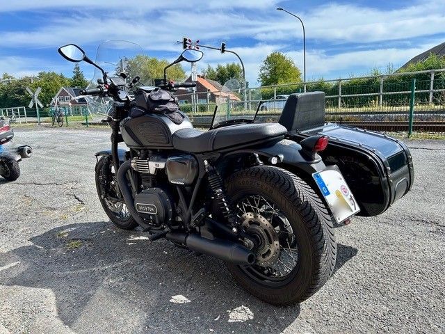 Schwarzes Motorrad mit Beiwagen, geparkt auf einem gepflasterten Parkplatz in der Nähe einer Bahnlinie.