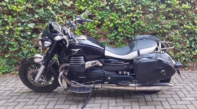 Schwarzes Moto Guzzi-Motorrad, geparkt auf einer gepflasterten Auffahrt vor einer grünen Hecke.