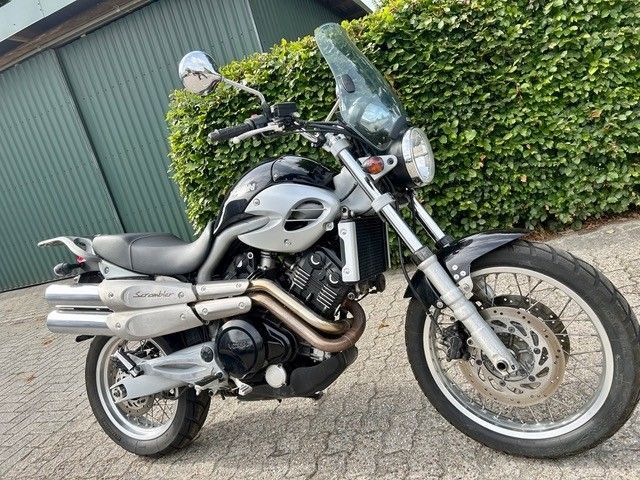 Motorrad parkt auf einer Straße, Seitenansicht, dahinter eine grüne Hecke und ein Wellblechzaun