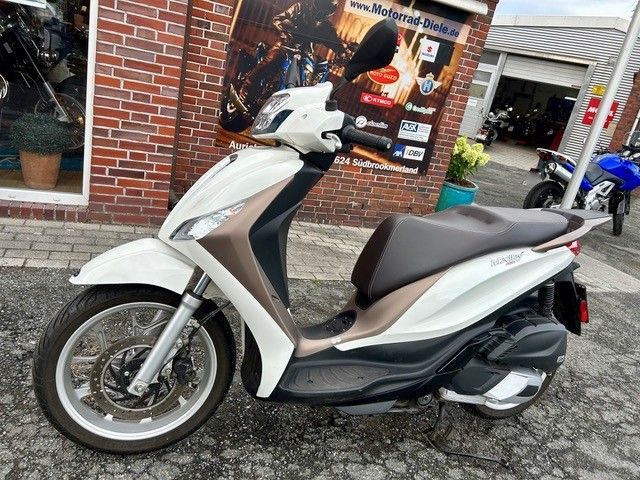 Ein weißer Roller parkt vor einem Backsteinladen mit Plakaten im Schaufenster.