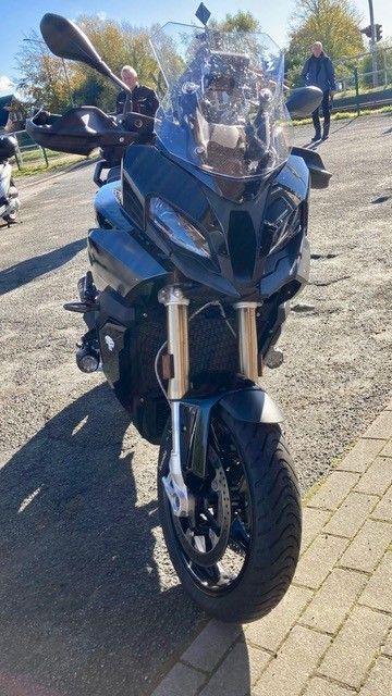 Schwarzes Motorrad auf gepflastertem Untergrund geparkt; im Hintergrund stehen zwei Personen.