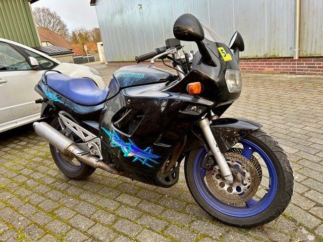 Auf einer gepflasterten Auffahrt steht neben einem silbernen Auto ein schwarz-blaues Motorrad.
