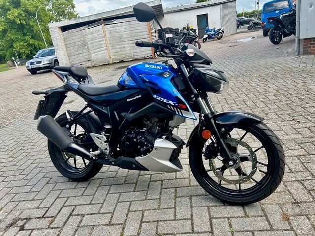 Ein blau-schwarzes Sportmotorrad parkt auf einem gepflasterten Parkplatz in der Nähe von Gebäuden und anderen Motorrädern.