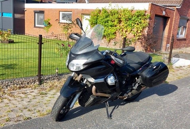 Schwarzes Moto Guzzi-Motorrad mit Seitentaschen auf einer Straße vor einem Backsteinhaus geparkt.