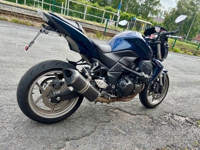 Ein blaues Sportmotorrad parkt auf einem rissigen Gehweg neben einem Metallzaun und Bäumen.