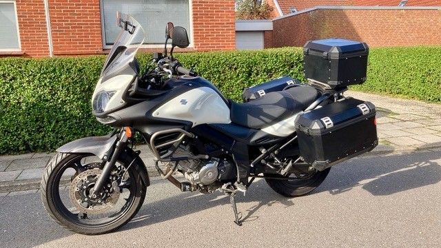 Silbernes Suzuki V-Strom-Motorrad mit schwarzem Gepäck. Draußen geparkt, mit grüner Hecke und Backsteingebäude im Hintergrund.