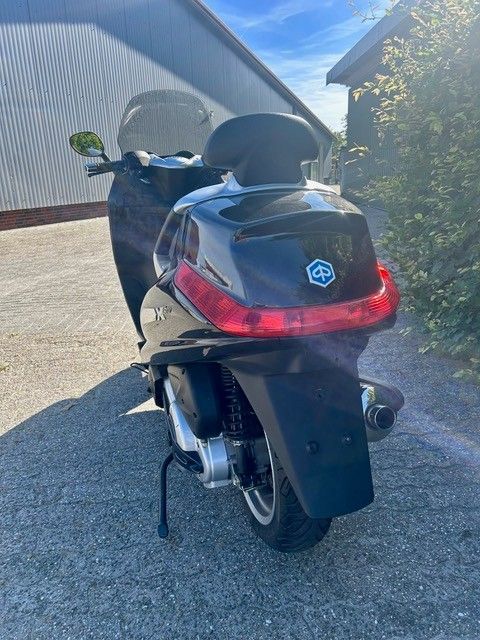 Schwarzer Roller parkt auf einer sonnigen Einfahrt neben einem Haus und einer Hecke, Rückansicht