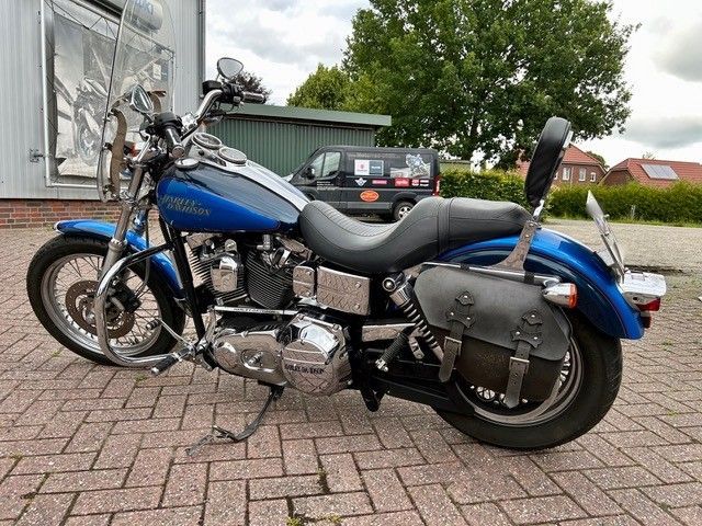 Blaues Harley-Davidson Dyna-Motorrad mit schwarzem Sitz, Satteltaschen und Chrom-Akzenten auf einer Ziegelfläche geparkt.