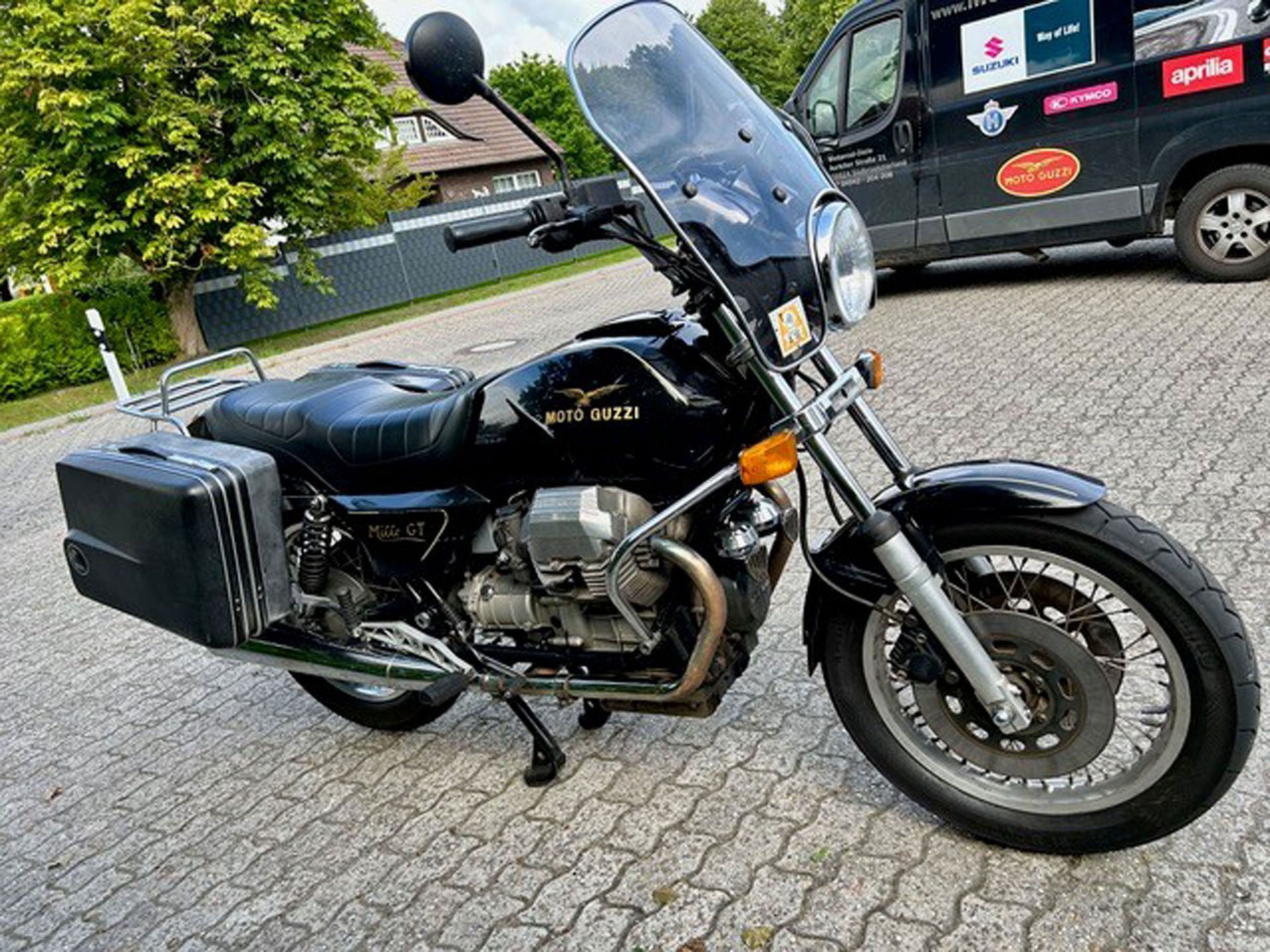 Schwarzes Moto Guzzi-Motorrad mit Windschutzscheibe, Seitengepäck und Chrom-Akzenten, geparkt auf Kopfsteinpflaster. Im Hintergrund ist ein schwarzer Lieferwagen zu sehen.