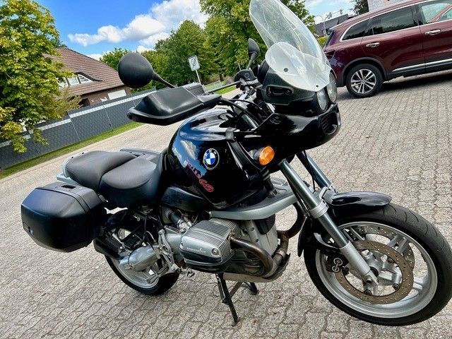 Schwarzes BMW-Motorrad mit Satteltaschen, geparkt auf einer Kopfsteinpflasterstraße, im Hintergrund ein Auto und ein Haus.