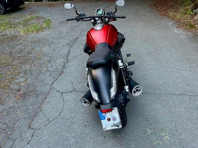 Ein rotes Motorrad parkt auf einer mit rissigem Schotter bedeckten Auffahrt, Ansicht von hinten.