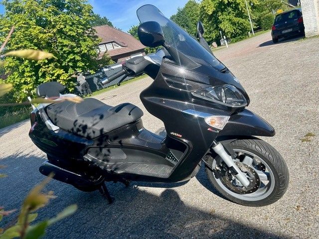 Ein schwarzer Roller parkt in einer sonnigen Wohnstraße mit Häusern und Bäumen im Hintergrund.