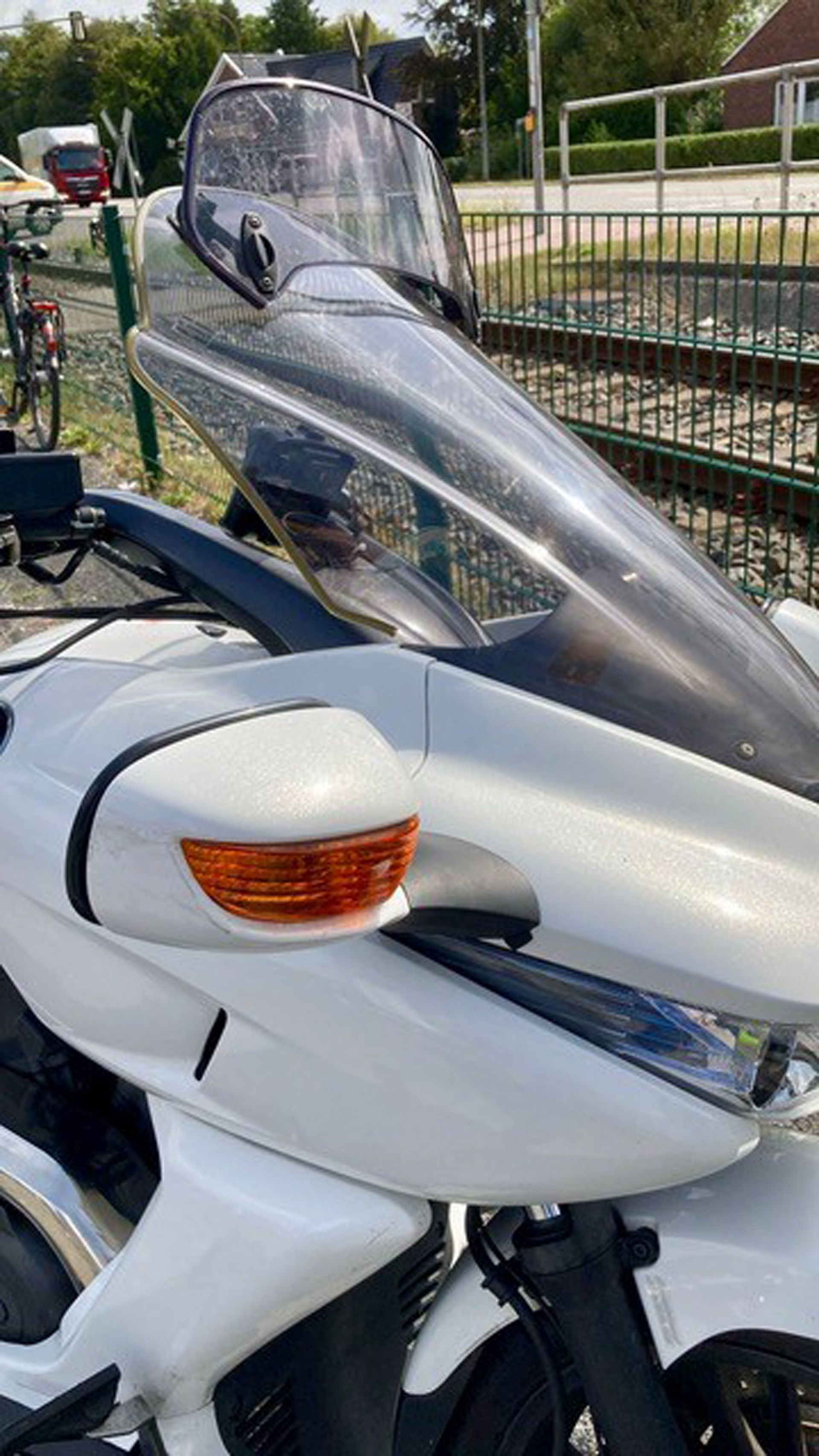 Weißes dreirädriges Motorrad mit großer Windschutzscheibe und orangefarbenem Blinker, draußen in der Nähe von Bahngleisen geparkt.