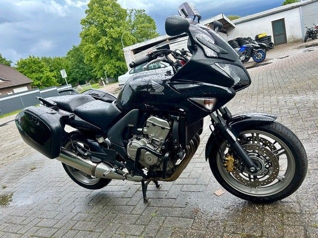 Ein schwarzes Tourenmotorrad steht auf einer nassen Ziegelsteinauffahrt; im Hintergrund sind ein weiteres Motorrad und eine Garage zu sehen.