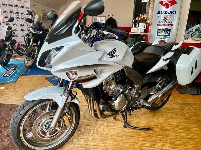 Ein silbernes Suzuki-Sportmotorrad ist in einem Ausstellungsraum zu sehen, im Hintergrund sind weitere Motorräder abgebildet.
