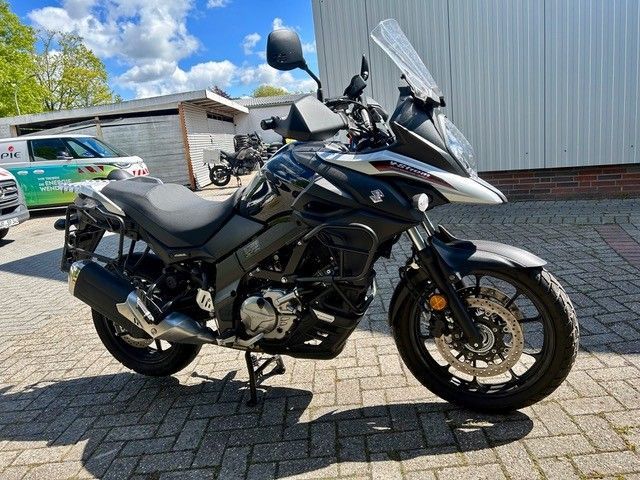 Schwarzes Adventure-Motorrad mit Windschutzscheibe und Topcase steht auf einem gepflasterten Parkplatz neben einem Gebäude.