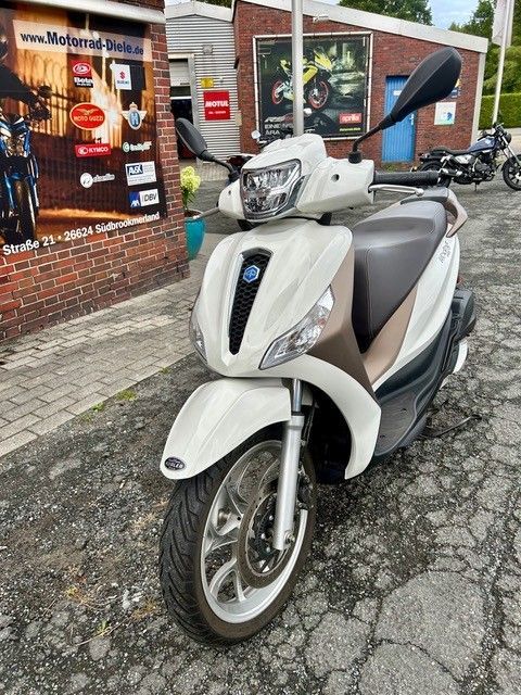 Ein weißer Roller parkte vor einem Backsteinladen mit Plakaten im Fenster auf nassem Gehweg.