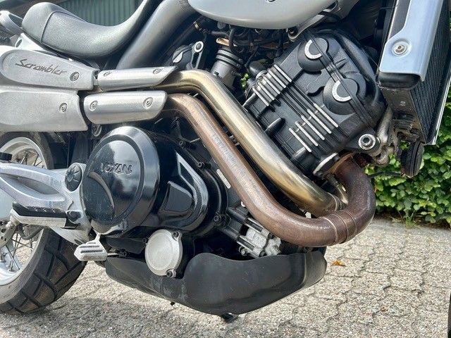 Nahaufnahme eines Motorradmotors und Auspuffrohren, die auf einem Gehweg in der Nähe von Grünflächen geparkt sind